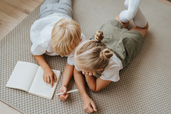 Birds-eye view of two children, lying on their fronts, working together in an exercise book